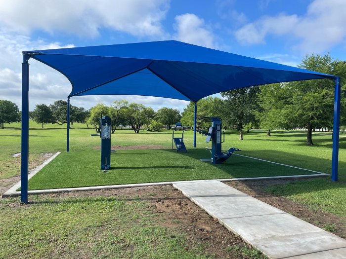 Outdoor fitness park with centralized outdoor fitness equipment installed beneath a shade canopy in a municipal park.