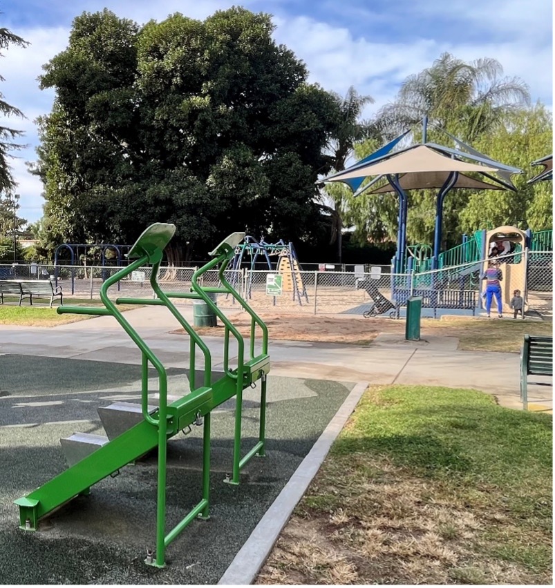 Outdoor fitness equipment installed near a park playground with clear sightlines for supervising parents
